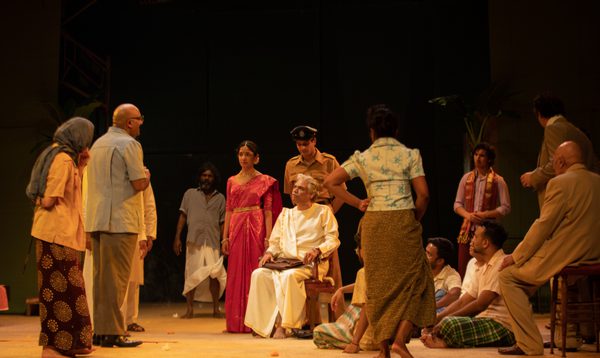 A man in white robes sits handcuffed to a chair with a policeman behind him and people gathered around him looking concerned.