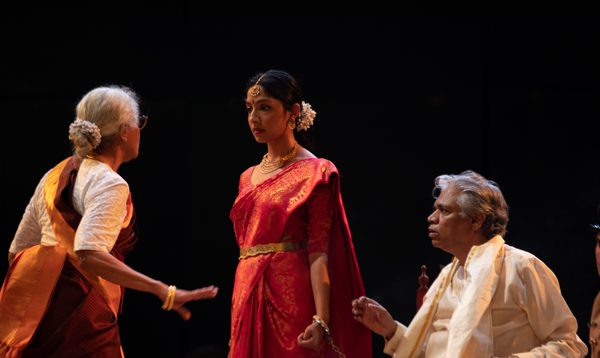 A woman in a red sari stands defiantly handcuffed to a chair with an old man next to her sat down in the chair, looking at an older woman.