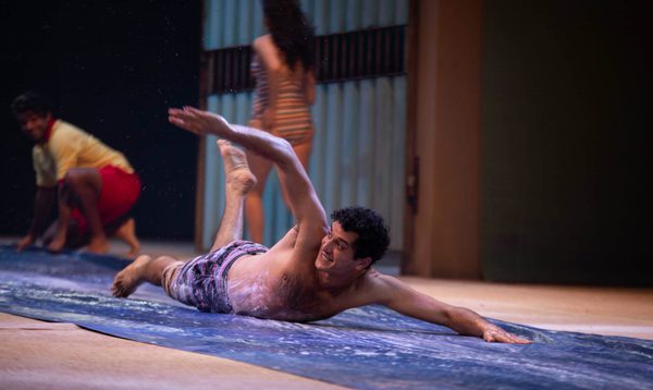 A man wearing swimming trunks slides on his front down a slip and slide with a grin on his face
