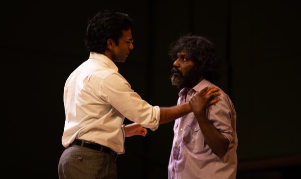 A well-groomed man in a white shirt and glasses stands with his hand on the shoulder of a man with overgrown hair and beard to comfort him.