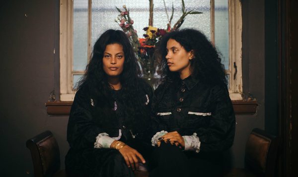 Two sisters sit side by side, one looking forward into the camera whilst the other gazes at her and a vase of flowers behind them