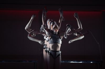 A line of dancers dressed as robots hold artificial limbs in a spider formation
