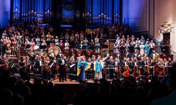 An orchestra stands on stage with two women at the front wrapped in Ukrainian flags holding hands