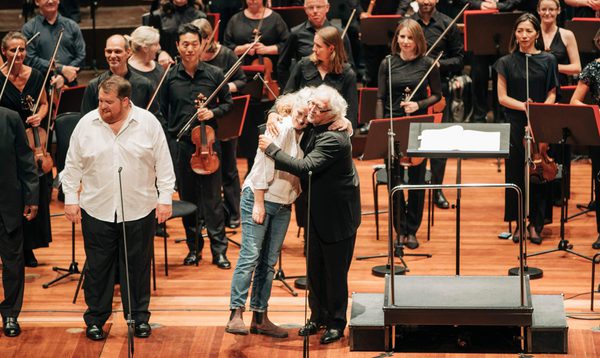 An orchestra stand on stage whilst the conductor and soloist hug at the front