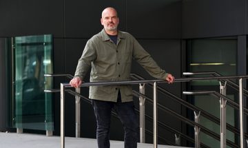 A man stands in a khaki shirt and blue jeans leaning on a railing looking into the camera.