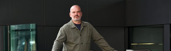 A man stands in a khaki shirt and blue jeans leaning on a railing looking into the camera.