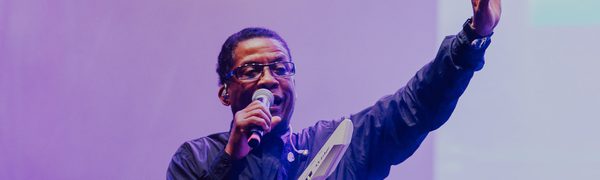 A man stands with a microphone in one hand and his other arm raised with a keytar around his neck in front of a purple background.