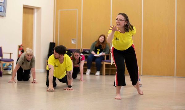 A woman in a yellow t-shirt leans forward with her hand in a paw shape, whilst three people crawl on the floor behind her