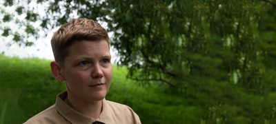 A artist pictured from the shoulders up, wearing a brown shirt and smiling slightly, standing outside with a tree and hedge in the background.