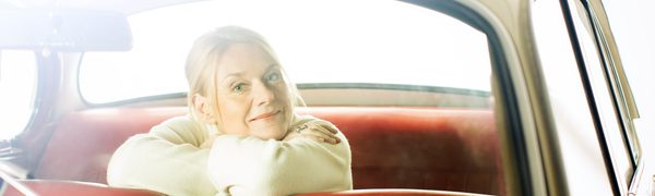 A woman sits inside a car with her head resting over her arms and looking out from the back of the car to the camera