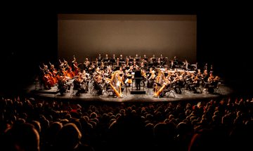 The orchestra shown onstage in the distance, with the silhouettes of rows of audience members in the foreground.