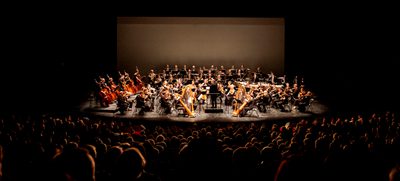 The orchestra shown onstage in the distance, with the silhouettes of rows of audience members in the foreground.