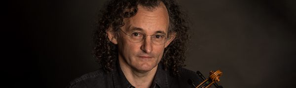 A man with shoulder-length curly hair, wearing a black shirt and wire-rimmed glasses, sits holding a violin, looking up at the camera.