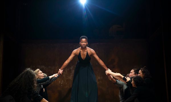 A woman with short afro hair in a black dress stands with her arms held either side by a group of women looking up at her