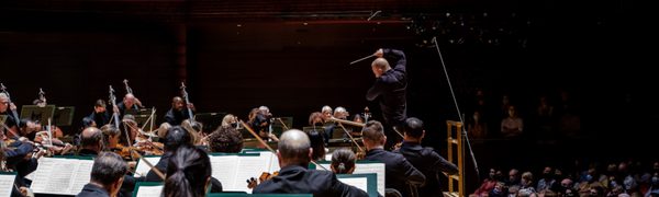 A conductor is standing with his right arm in the air in front of a full orchestra on a stage, with an audience looking on.
