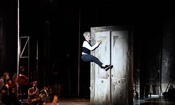 A man in a waistcoat and suit trousers is seen clinging to a door frame