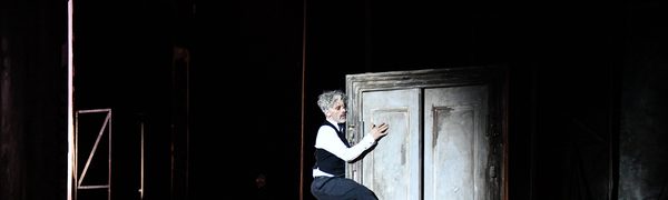 A man in a waistcoat and suit trousers is seen clinging to a door frame