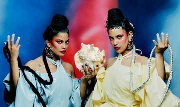 Two woman in matching puffy dresses, one blue and one yellow, sit holding a conch shell in one hand and their braids in the other.