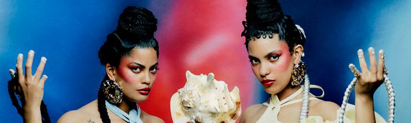 Two woman in matching puffy dresses, one blue and one yellow, sit holding a conch shell in one hand and their braids in the other.