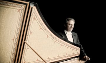 Richard Egarr dressed in a suit and a white bow tie stands behind a harpsichord with the lid open.