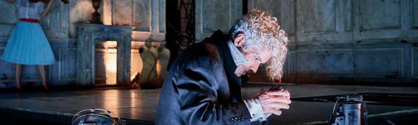 A man with curly white hair sits on the floor leaning over a contraption of some sort, with a woman in a white dress playing the violin in the background.