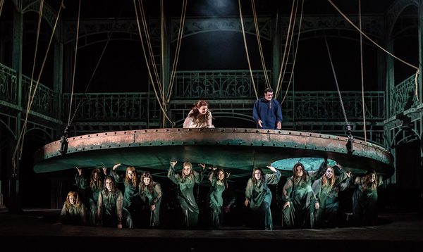 A man and woman stand above a raised disk smiling, with women dressed as water nymphs stood below the disk looking fierce.