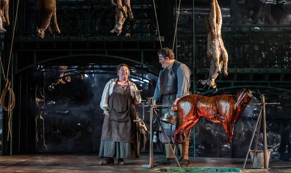 A man and a woman dressed in butcher's clothing stand on stage with a roast pig and dead pigs hanging from the ceiling.