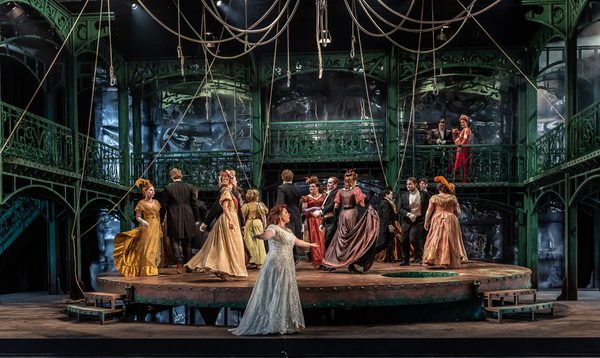 People in extravagant ball dress stand and dance on a raised circular platform on stage
