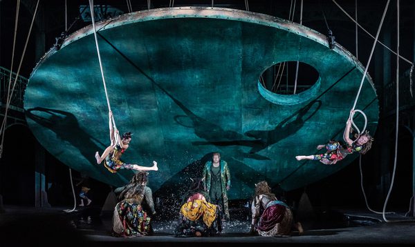 Singers in folk-style costume are on stage, splashing in a large pool of water, with two acrobats swinging in front.