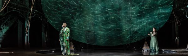 A man and a woman stand on stage under a large suspended disk with watery projections behind them.