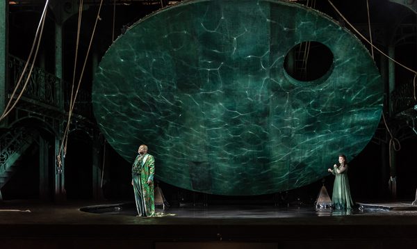 A man and a woman stand on stage under a large suspended disk with watery projections behind them.
