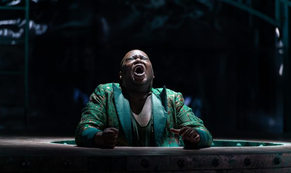 A man stands singing through a hole in the stage with his fists clenched and his eyes shut.