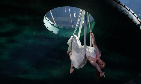 A woman in a white dress is suspended from above through a hole in the roof, leaning back and extending her arm.