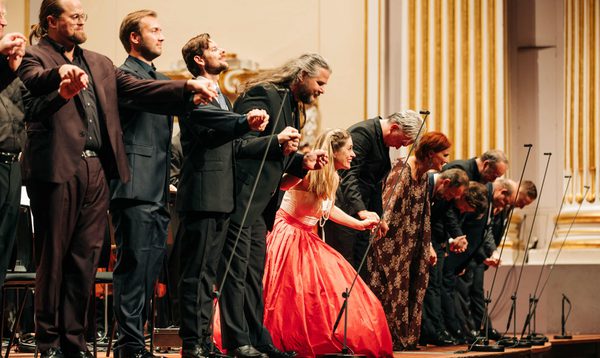 A group of men in suits and a woman in a vibrant pink dress bow and hold hands at the front of a stage