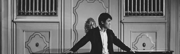A greyscale photo of Seong Jin Cho dressed in a black suit leaning against a grand piano with his arms stretched out to both side.