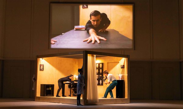 A man is seen on a screen reaching over a table above a box in which the same man stands opposite a camera operator