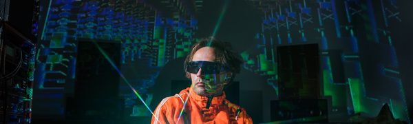 A man wearing an orange jacket and sunglasses holds a mirror in front of him, refracting blue and green rays of light around the room.