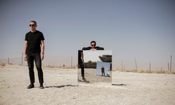 Two men stand in a desert, one of them leaning on a large mirror. You can see a reflection of them in another mirror.