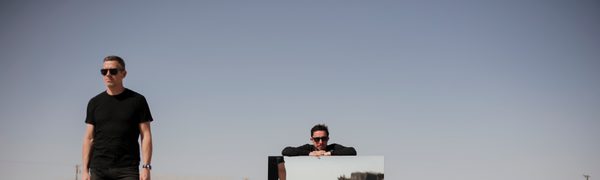 Two men stand in a desert, one of them leaning on a large mirror. You can see a reflection of them in another mirror.