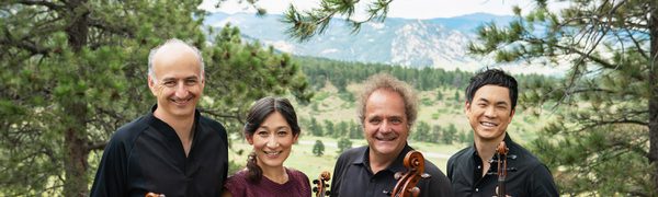 A string quartet stand in a grassy field against a mountainous backdrop, smiling towards the camera and holding their instruments.