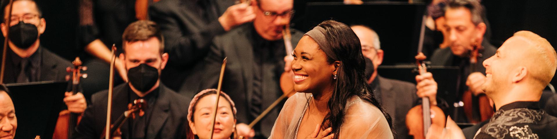 Angel Blue standing in front of an orchestra holding her hand to her chest and smiling