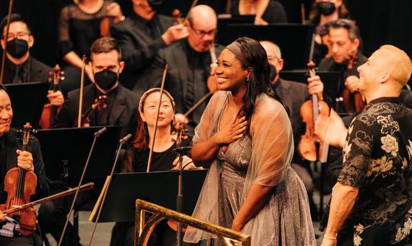 Angel Blue standing in front of an orchestra holding her hand to her chest and smiling
