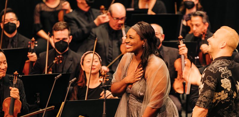 Angel Blue standing in front of an orchestra holding her hand to her chest and smiling