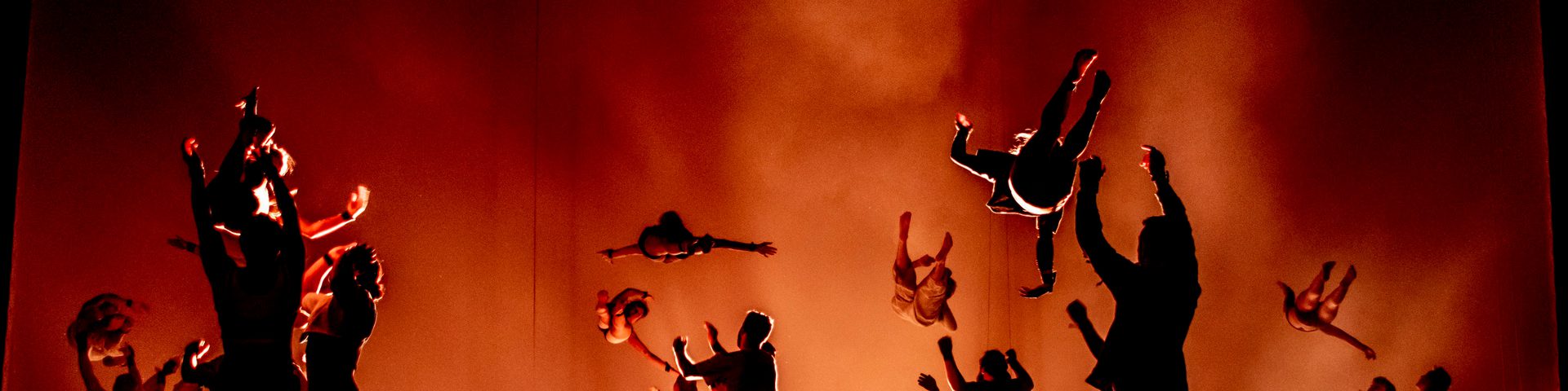 Lots of gymnasts are performing jumps and tumbles in silhouette against a red, smoky background.