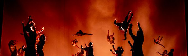 Lots of gymnasts are performing jumps and tumbles in silhouette against a red, smoky background.