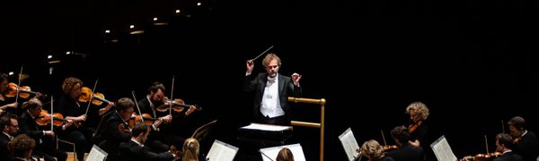 A conductor with his arms aloft in front of a sea of string instrument musicians
