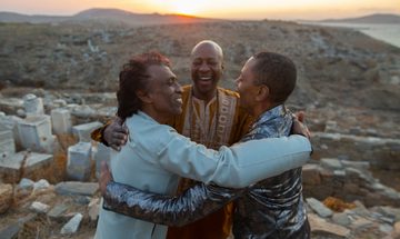 Three men stand in a desert landscape embracing and smiling at each other