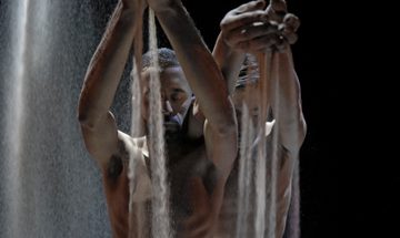 Two dancers on a spot-lit stage hold their arms in front of them as sand pours out of their hands.
