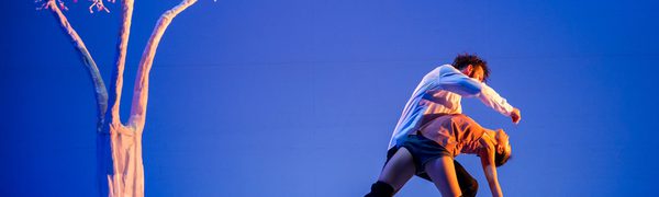 A man and a woman are dancing in a blue room with a cardboard tree.