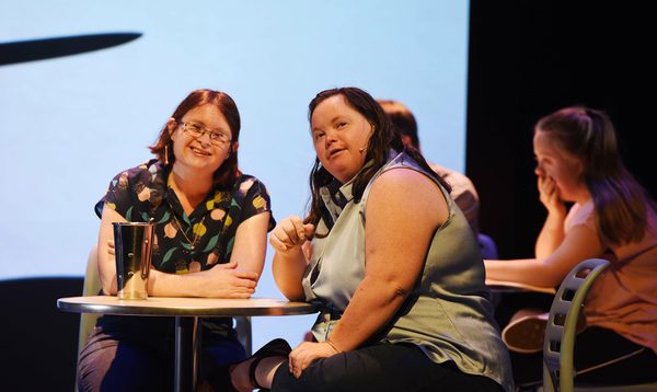 Two women with Down's syndrome sit at a diner table looking at something in the distance and smiling.